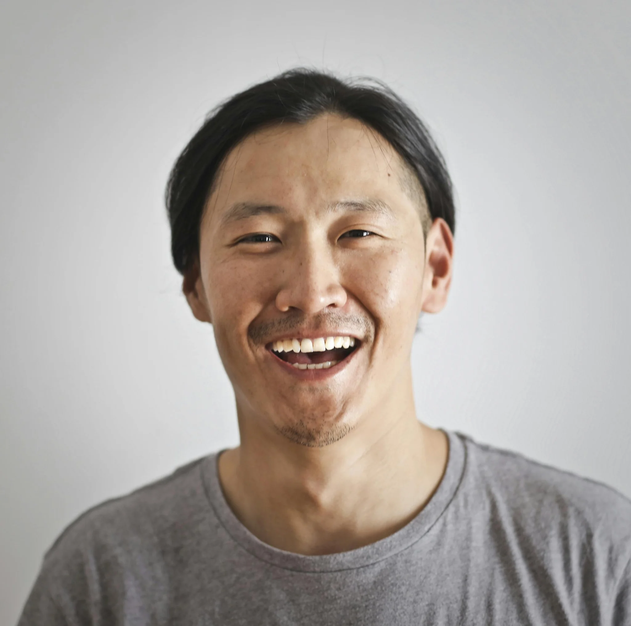 Smiling Asian man with dark hair in a gray shirt, showing a big grin with open mouth. 