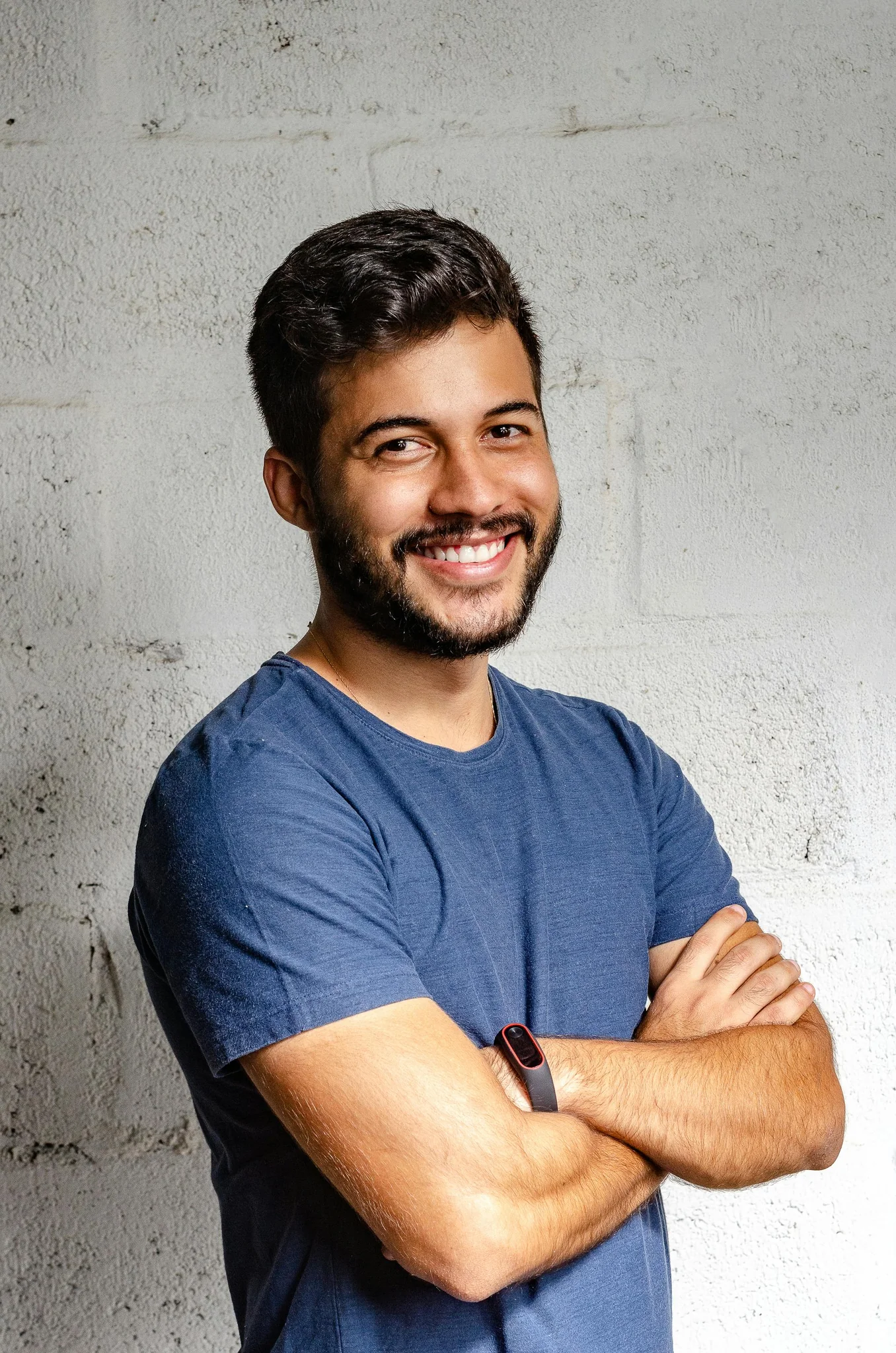 A smiling man with short dark hair stands with arms crossed, wearing a blue t-shirt against a neutral background.