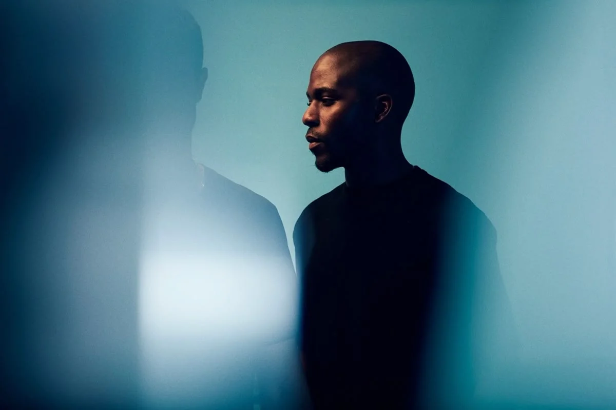 Portrait of a Black man with a shaved head in profile, looking contemplative against a blue backdrop with blurred foreground.