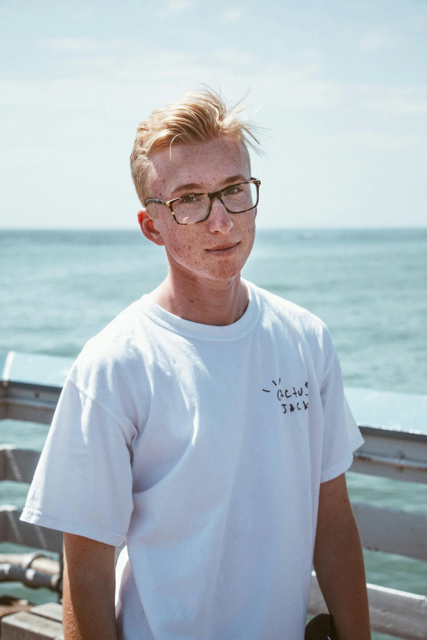 A young man with blond hair wearing glasses and a white t-shirt smiles at the camera on a pier.